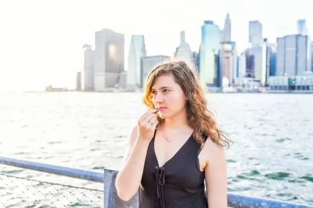 Young woman outside outdoors in NYC New York City Brooklyn Bridge Park by east river, railing, view of cityscape skyline, putting on lipstick makeup