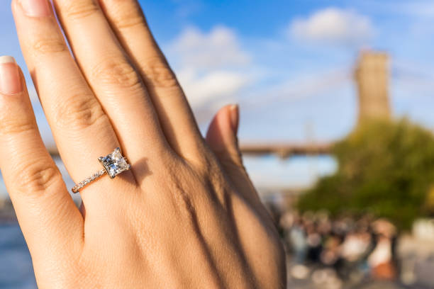 young woman's hand with diamond engagement ring princess cut, gold outside outdoors in nyc new york city brooklyn bridge park by east river, cityscape, skyline bokeh - brooklyn bridge bridge brooklyn stone imagens e fotografias de stock