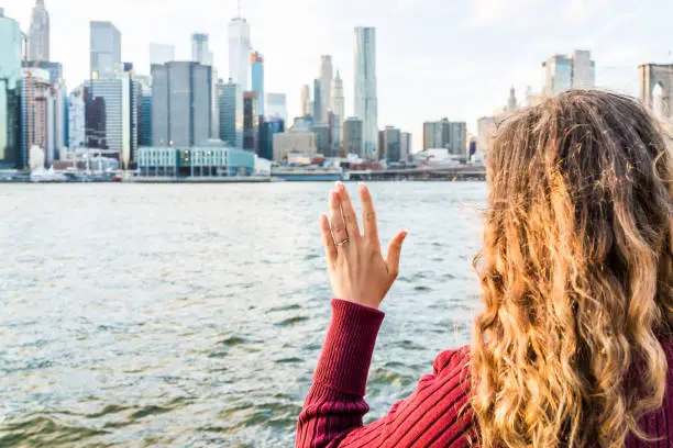 Young woman's hand with diamond engagement ring solitaire, gold wedding band outside outdoors in NYC New York City Brooklyn Bridge Park by east river, cityscape, skyline waving goodbye