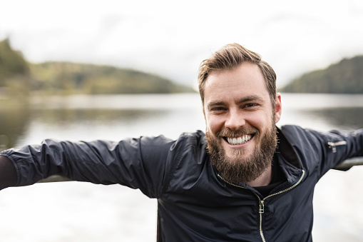 Bearded man sitting down outdoors in front of a lake on a moody day. Wearing a blue jacket looking at camera.
