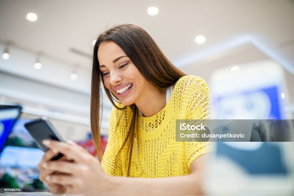 Close up view of satisfied young smiling attractive brunette girl testing mobile in a tech store. New Stock Photo