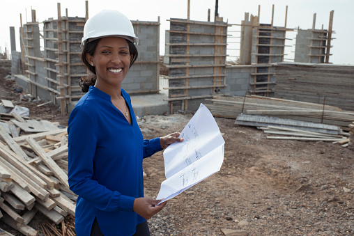 Woman civil engineer / architect holding blueprint of a house under construction, standing at the site.