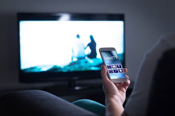 携帯電話でスマート tv リモート コントロール アプリケーションを使用してテレビを見ている男。 - home movie ストックフォトと画像