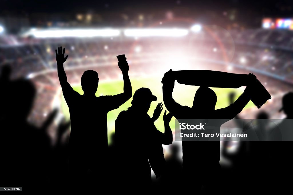 Friends at football game in soccer stadium. Crowd cheering and celebrating a goal in arena during match. Friends at football game in soccer stadium. Crowd cheering and celebrating a goal in arena during match. Silhouette people in live sport audience having fun. Soccer Stock Photo