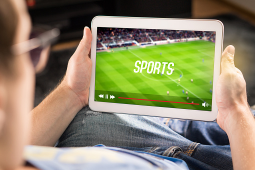 Man watching sports on tablet. Football and soccer game live stream and video player on screen. Pay per view (PPV) service. Replay or highlights broadcast. Lazy person relaxing.