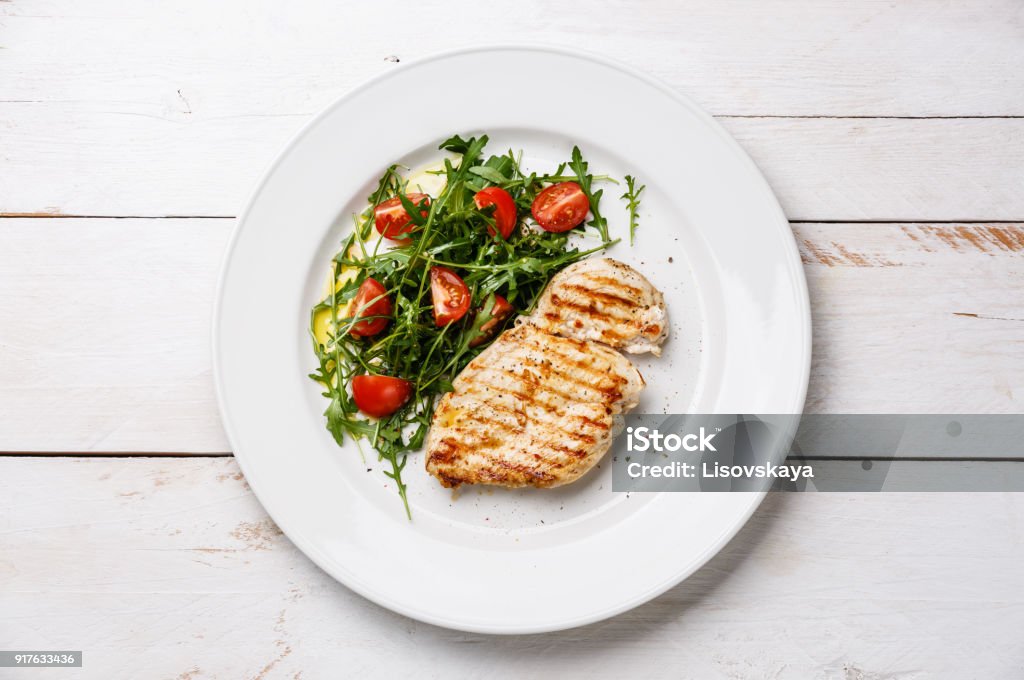 Roasted grilled chicken breast and fresh salad Roasted grilled chicken breast and fresh salad with tomato and arugula on wooden background Plate Stock Photo