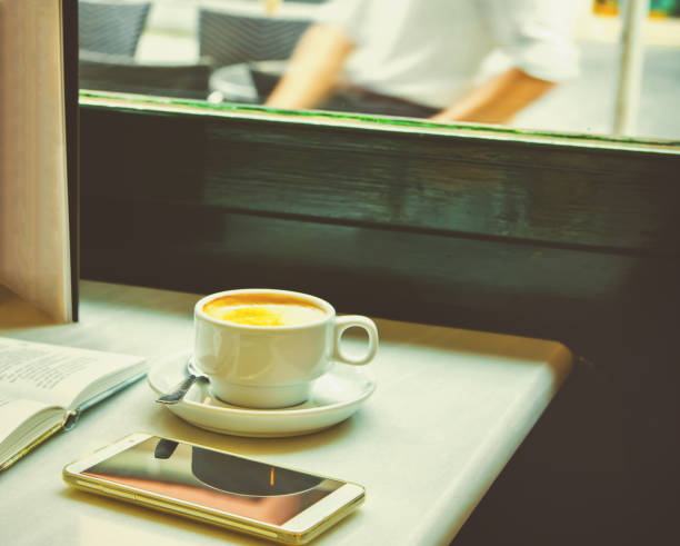 A close-up image of a cup of coffee on wooden table, with smartphone, notepad, pen and eyeglasses in the background, copy space.