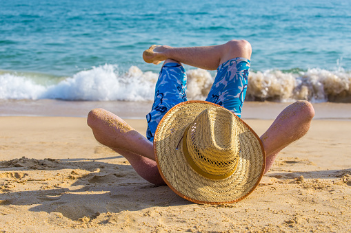 Young caucasian man with hat lying on beach at sea. My son was enjoying the sun while he was relaxing on the sand near the ocean. Water waves are coming from the blue water towards him. Sunbathing and lying lazy in nature is fun to do when you are on vacation in Portugal.