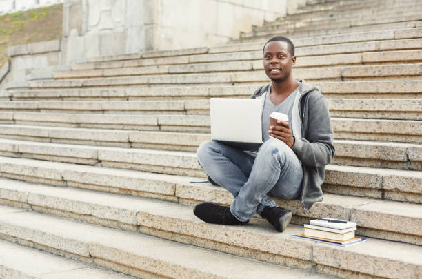 istock Smiling african-american student sitting on stairs with coffee using laptop 917656302
