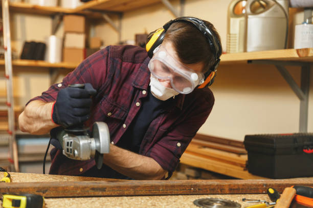Caucasian young man in plaid shirt, black T-shirt, noise insulated headphones, protective mask working in carpentry workshop at wooden table place with different tools, sawing iron with power saw joven caucásica en auriculares de aislamiento de ruido de camisa a cuadros, camiseta negra, máscara protectora trabajando en taller de carpintería en el lugar de la mesa de madera con diferentes herramientas, cortar hierro con poder vio.