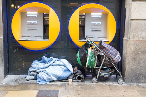BARCELONA, SPAIN, February 4, 2018 Homeless man covered with blankets sleeping under ATMs, near him stands a stroller with his stuff