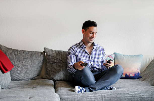 Smiling man on couch using remote control and smartphone stock photo