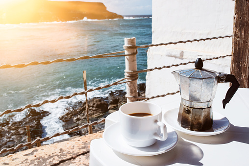 moka pot espresso maker and cup of coffee on table with coastline and ocean in background on sunny day