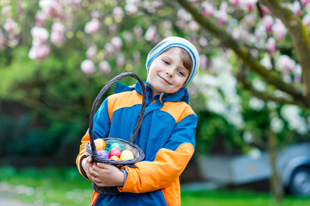 Cute adorable little kid boy making an egg hunt on Easter. Happy child searching and finding colorful eggs in domestic garden. Boy in spring clothes on cold day. Old christian and catholoc tradition