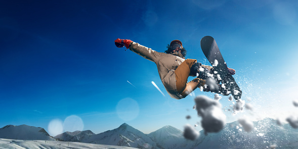 Male extreme snowboarder jumps and do a spectacular stunt in the air against a background of blue skies and snowy mountains. Freestyle snowboarding.