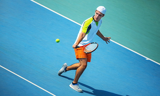Closeup side view of a young man playing tennis on a sunny summer day. He's hitting single hand forehand against blue hard court. He's wearing tennis clothes and a white cap pointed backwards.
