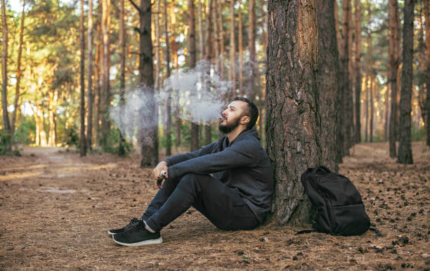 istock Man vaping outdoor. bearded young man having rest and sitting under the tree in forest. 917759228