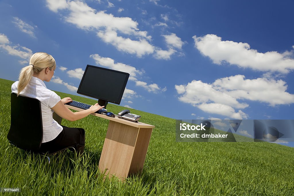 Businesswoman at desk using computer in green grass field Business concept shot of a beautiful young woman sitting at a desk using a computer in a green field with a bright blue sky with fluffy white clouds. Shot on location. 20-29 Years Stock Photo