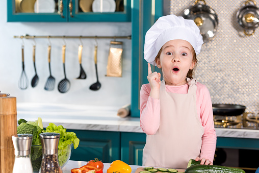 excited little child in chef hat and apron pointing up with finger and looking at camera in kitchen