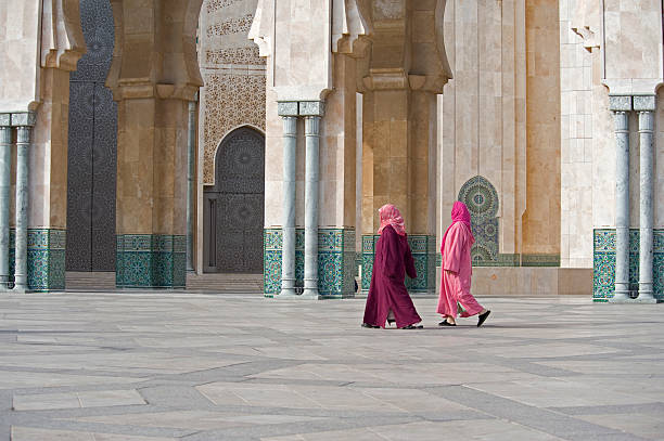 Two women walk in Hasan II Mosque in Casablanca, Morocco Two figures walking along the many spatial gates in wrought copper that give entrance to the Hassan II Mosque in Casablanca, the second largest religious building in the world(after Mekka) with room for 25 000 worshippers inside. casablanca morocco stock pictures, royalty-free photos & images