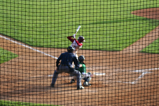 Baseball hitter waits for the next pitch - with catcher and umpire set for action. Deliberate shallow depth of field with players blurred beyond a protective net screen.