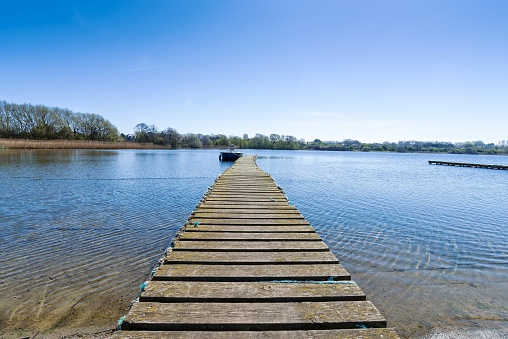 Wooden planked walkway stretches out into the lake with a moored boat at its end. Trees line the edge of the lake.