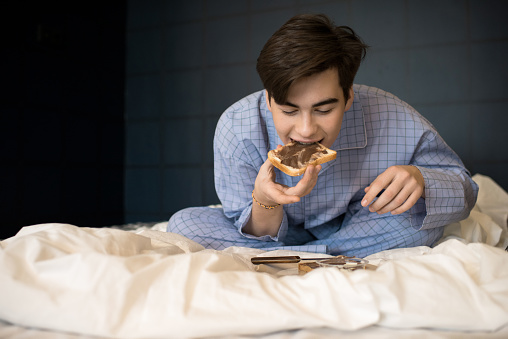 Portrait of teenage boy eating in bed having late night snack of toast and chocolate paste, copy space