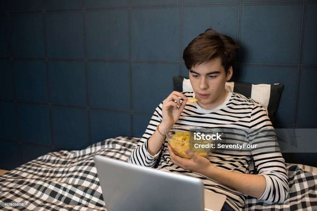 Teenage Boy on Weekend Night Portrait of teenage boy using laptop in bed and eating chips while watching movies at home, copy space Teenager Stock Photo