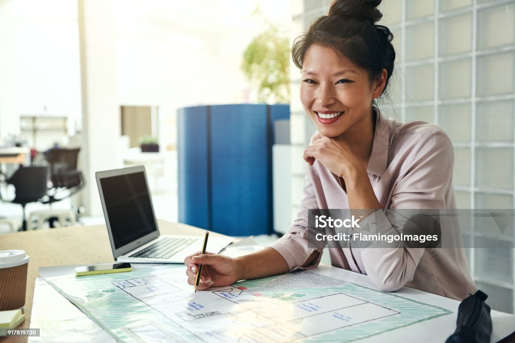 Smiling Asian architect working on blueprints at an office desk Smiling young Asian architect sitting alone at her desk in a modern office working on a building design Architect Stock Photo