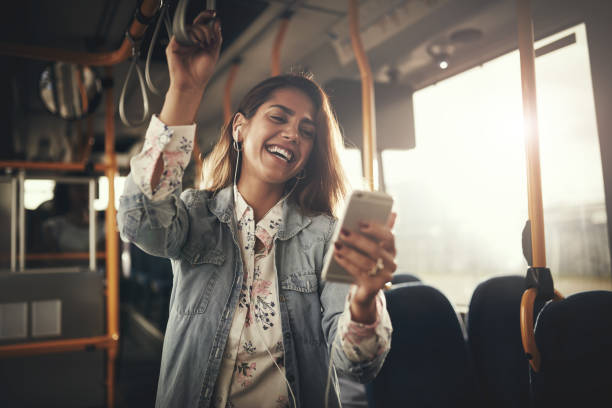 Young woman laughing while listening to music on a bus Young woman wearing earphones laughing at a text message on her cellphone while riding on a bus public transportation stock pictures, royalty-free photos & images