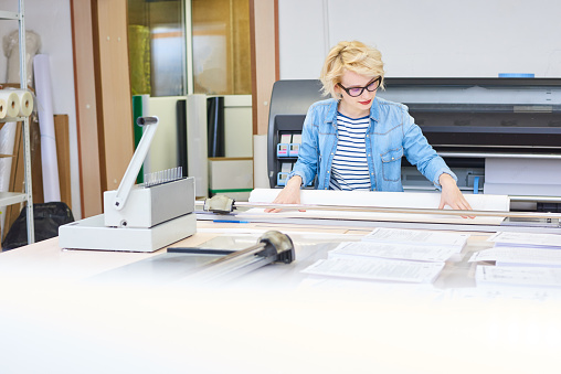 Portrait of blonde young woman working in modern printing shop or publishing company, cutting  paper and loading plotter machines, copy space