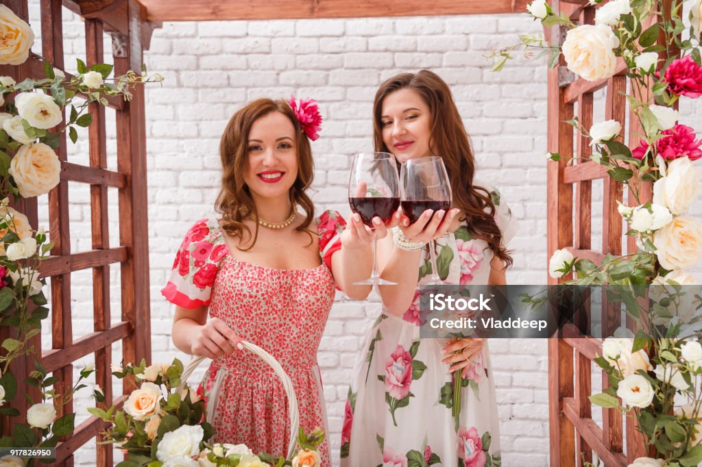 Dos Chicas Guapas Vestidas Con Vestidos De Verano Posando Junto A Un Arco De Flores Con Vasos De Vino Tinto En Las Manos Foto de stock y más banco de imágenes de Dos Chicas Guapas Vestidas Con Vestidos De Verano Posando Junto A Un Arco De Flores Con Vasos De Vino Tinto En Las Manos Foto de stock y más banco de imágenes de