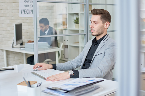 Side view portrait of young businessman working at desk using computer in modern office, copy space