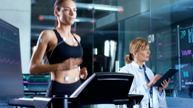 Beautiful Woman Athlete Runs on a Treadmill with Electrodes Attached to Her Body, Female Physician Uses Tablet Computer and Controls EKG Data Showing on Laboratory Monitors.