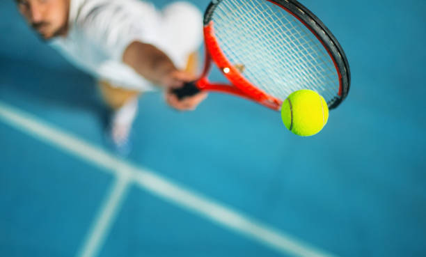 Tennis game at night. Closeup high angle view of two male tennis player competing in an outdoors night match. He's hitting a serve or a volley against blue hard court with copy space. tennis ball stock pictures, royalty-free photos & images