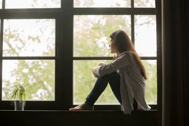 Photo of Thoughtful girl sitting on sill embracing knees looking at window