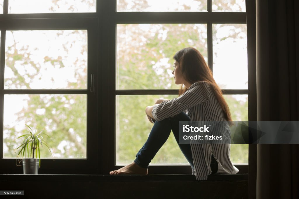 Menina pensativa, sentada no peitoril, abraçando os joelhos, olhando para a janela - Foto de stock de Mulheres royalty-free