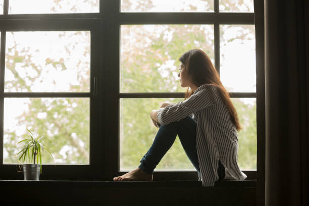 thoughtful girl sitting on sill embracing knees looking at window - depression women sadness window imagens e fotografias de stock