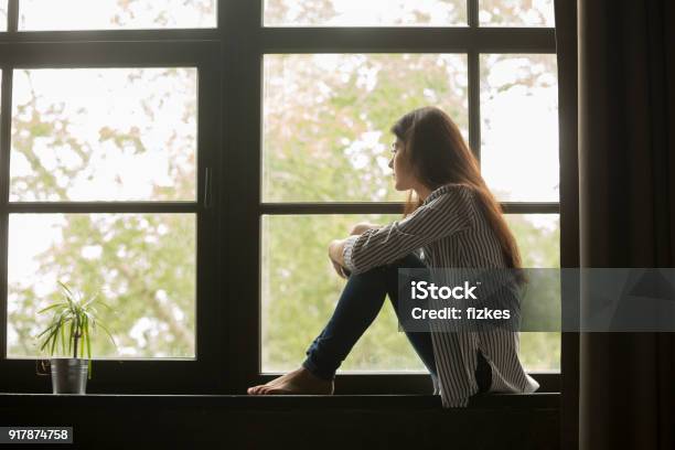 Foto de Menina Pensativa Sentada No Peitoril Abraçando Os Joelhos Olhando Para A Janela e mais fotos de stock de Mulheres