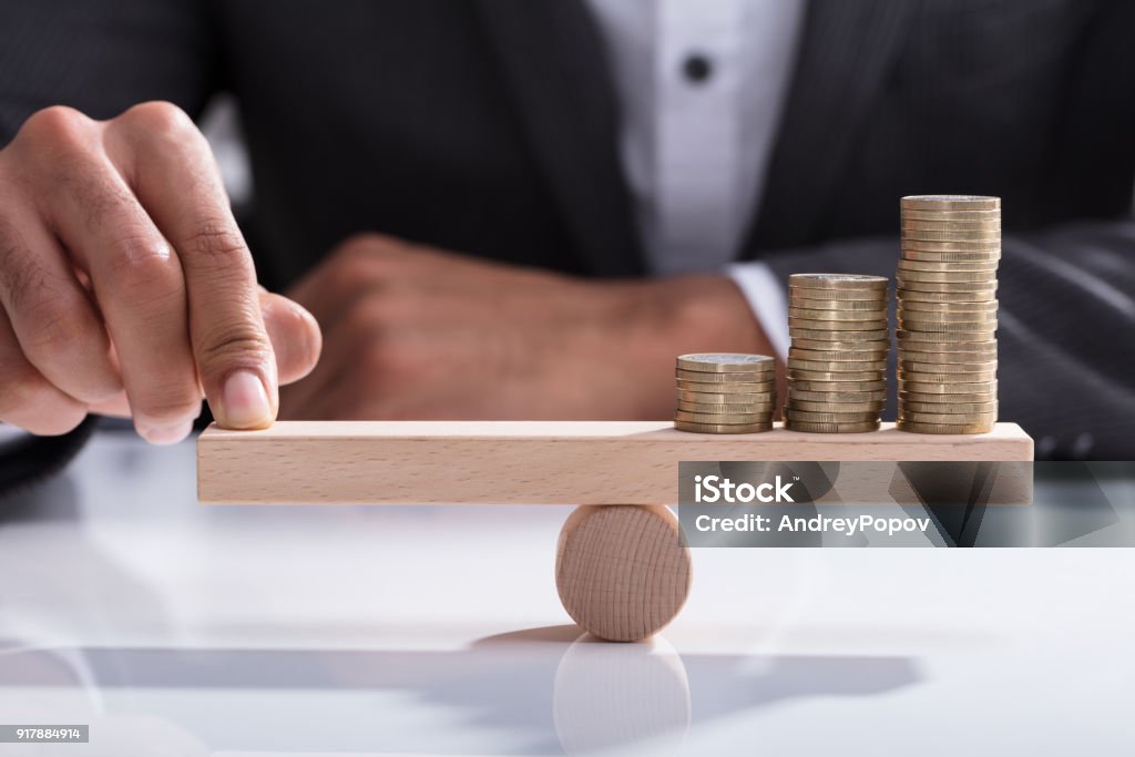 Businessperson Balancing Stacked Coins On Wooden Seesaw Close-up Of A Businessperson's Hand Balancing Stacked Coins On Wooden Seesaw With Finger Over Desk Balance Stock Photo