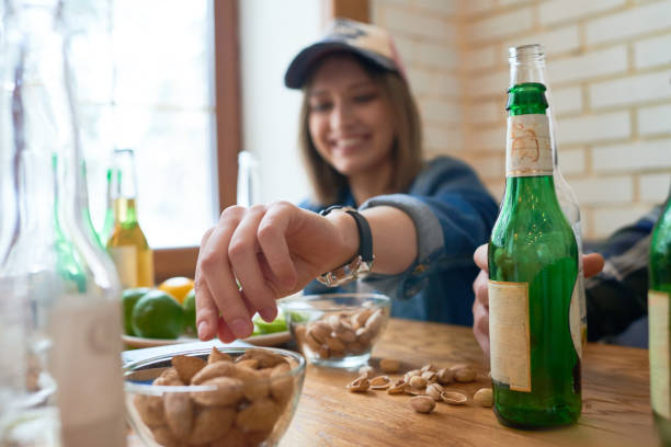 Close-up of jolly tomboy eating nuts while drinking beer while sitting at table with friends in modern bar