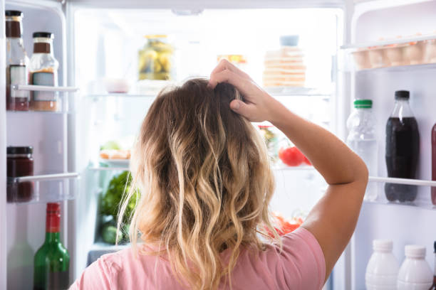 混乱女性オープン冷蔵庫の中を見ています。 - hungry ストックフォトと画像