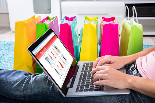 Woman Sitting On Carpet Using Laptop To Shop Online Besides Colorful Shopping Bags