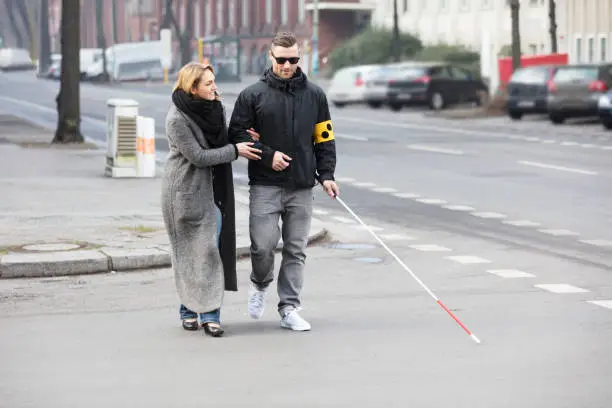 Young Woman Assisting Blind Man With White Stick On Street