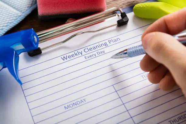 Close-up Of Person Hand Filling Weekly Cleaning Plan Form With Pen
