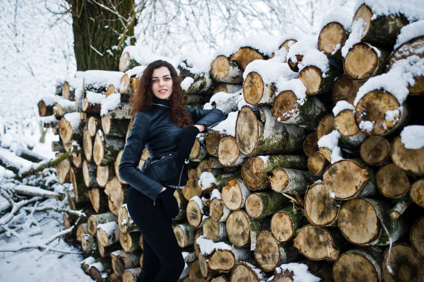 Elegance curly girl in leather jacket at snowy forest park at winter against stumps.