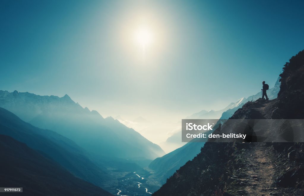 Standing woman on the hill against mountain valley at bright sunny day. Landscape with girl, trail, mountain, blue sky with sun and low clouds at sunset in Nepal.  Lifestyle, travel. Trekking Mountain Stock Photo