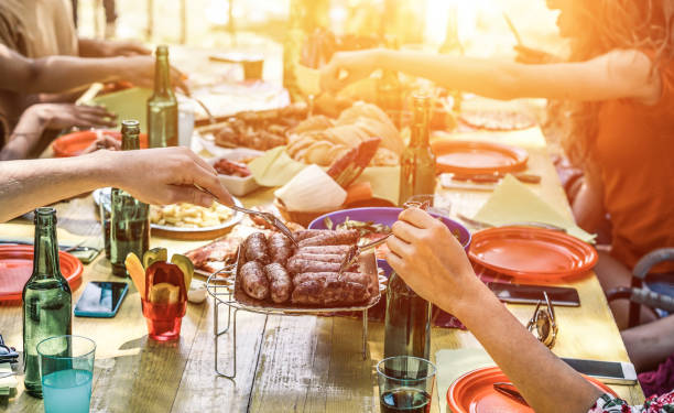 group of happy friends eating and drinking beers at barbecue dinner at sunset - adult people having meal together outdoor - focus on fork sausages - summer lifestyle, food and friendship concept - picnic family barbecue social gathering imagens e fotografias de stock
