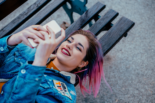 Smiling girl with purple hair lying on a bench, using phone.