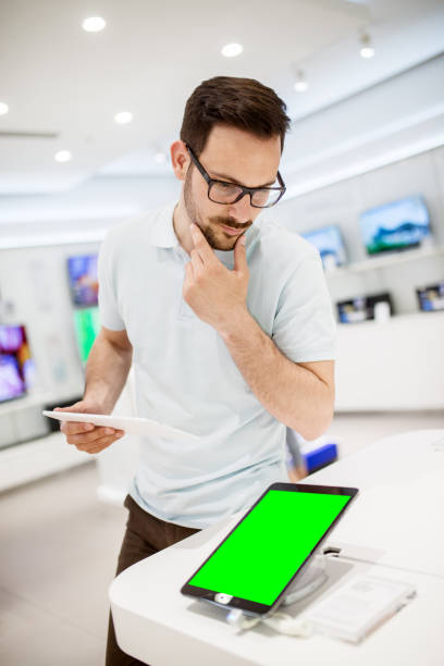 istock Curious young bearded handsome man looking and thinking about the new tablet in a tech store with the blank editable green screen. 917930362